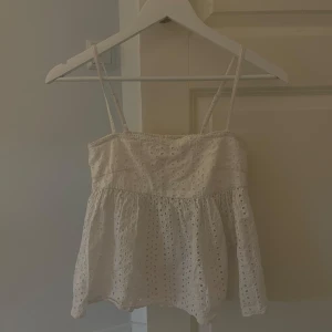 Vit broderad linnetopp med spets - Supersöt vit linnetopp med tunna axelband och broderade hål i tyget. Toppen har en lätt volang nertill och fina spetsdetaljer! Perfekt när man vill ha en luftig och söt topp till varma tillfällen!☀️