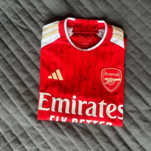 Arsenal Saka 7 matchtröja Adidas XS - Röd och vit Arsenal fotbollströja från Adidas med Saka 7-tryck på ryggen. Tröjan har blixtmönster, guldiga Adidas-detaljer och klubbmärke på bröstet. Kortärmad modell i lätt funktionsmaterial, perfekt för match eller träning.