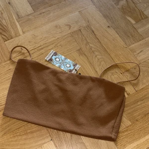 Brun bandeau topp från Boohoo - Snygg brun bandeau topp från Boohoo med tunna axelband. Toppen har en enkel och stilren design i ett mjukt, lätt strukturerat material. Perfekt att matcha med höga jeans eller kjol för en trendig look.
