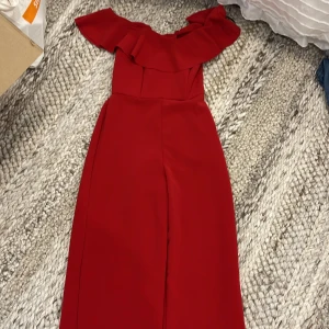 Röd offshoulder jumpsuit H&M - Snygg röd jumpsuit från H&M med offshoulder och volang upptill. Har en figurnära passform och dragkedja i ryggen för enkel påklädning. 
