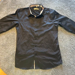 Svart Burberry skjorta med rutiga detaljer - Snygg svart skjorta från Burberry i storlek M. Klassisk modell med långa ärmar och knappar framtill. Krage och ärmslut har Burberrys ikoniska rutiga mönster i beige, svart, vitt och rött. Perfekt för dig som vill ha en stilren look med exklusiv känsla.