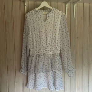 Vit blommig kortklänning Gina Tricot - Supersöt vit kortklänning från Gina Tricot med små blå blommor, långärmad och med volangdetaljer😍. Klänningen har smock i midjan och är gjord i ett lätt, luftigt material!💓 säljer pgv den inte kom till användning men är är bra skick och inga hål, fläckar osv!💞