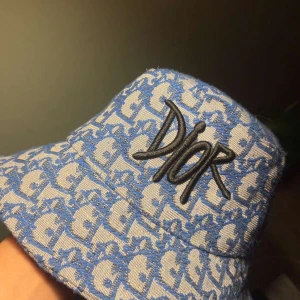 Blå bucket hat från Dior - Jag säljer en stilig bucket hat från Dior i blått och vitt med ikoniskt Dior-mönster över hela hatten. Broderad Dior-logga i svart framtill. Jag säljer den för den inte kommer till så mycket användning. Priset går att diskuteras vid snabb affär