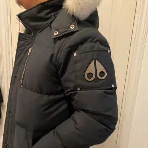 Svart dunjacka Moose Knuckles päls - Snygg svart dunjacka från Moose Knuckles med fluffig vit pälskrage. Jackan har coola metalldetaljer på ärmen, dragkedja framtill och bröstficka med dragkedja. Perfekt för kalla dagar och riktigt street i stilen. Det är storlek L passar även storlek M .Jag själv håller på flyttar och vill gärna bli av med den så snabbt som möjligt men om de kommer ett snabbt bud så kan det tas för ett good pris 