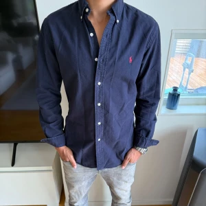 Marinblå skjorta från Ralph Lauren - Klassisk marinblå skjorta från Ralph Lauren med knappar hela vägen och ikonisk röd logga broderad på bröstet. Skjortan har lång ärm, button-down krage och är tillverkad i mjuk bomull. Perfekt för en stilren och avslappnad look.