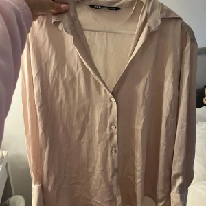 Beige silkesblus från Zara - Säljer en stilren beige skjorta från Zara i glansigt silkesmaterial. Skjortan har klassisk krage, knappar framtill och långa ärmar. Perfekt för dig som gillar en clean och elegant look.