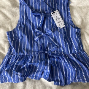 Blå randig peplumtop från Gina Tricot - Superfin blå peplumtop från Gina Tricot med vita ränder och knytband framtill. Toppen är ärmlös och har en volangkant nedtill som ger en söt och trendig look. Perfekt till jeans eller kjol för en fräsch sommarstil.