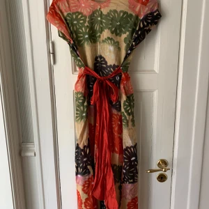 Blommig långklänning med rött knytband - Färgglad långklänning med stora blommor i rött, grönt, svart och rosa på beige botten. Klänningen har v-ringning, korta ärmar och ett rött knytband i midjan som ger en snygg siluett. Perfekt för sommarens alla tillfällen.