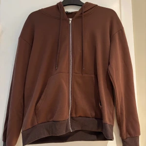 Brun hoodie med dragkedja från SHEIN - Mysig brun hoodie från SHEIN i storlek S. Tröjan har huva med snörning, hel dragkedja framtill och två stora fickor. Tillverkad i mjukt fleecematerial som är perfekt för kyliga dagar. Långärmad och med ribbade muddar vid ärmslut och nederkant.