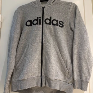 Grå hoodie från Adidas med dragkedja - Säljer en grå hoodie från Adidas med svart logga framtill och dragkedja hela vägen. Tröjan har huva, två fickor och ribbade muddar. Perfekt för chill dagar eller till träningen. Klassisk och enkel stil som funkar till det mesta.