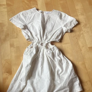 Vit cut out klänning från Zara - Superfin vit kortklänning från Zara med coola cut out-detaljer i midjan och rund halsringning. Klänningen har korta ärmar och är gjord i bomull, perfekt för varma dagar. Snygg och trendig modell som verkligen sticker ut!