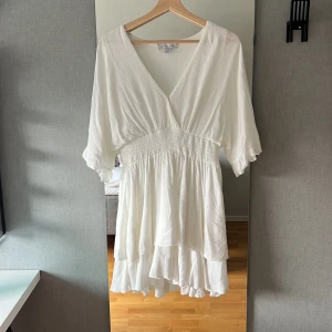 Vit kortklänning från Amisu - Superfin vit kortklänning från Amisu med v-ringning, smockad midja och volangig kjol. Klänningen har korta ärmar med lätt puff och är tillverkad i ett tunt, luftigt tyg som ger en skön känsla. Perfekt för varma dagar och somriga vibes!