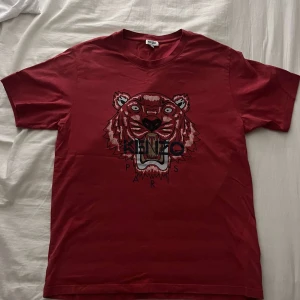 Röd Kenzo t-shirt med tigertryck - Snygg röd t-shirt från Kenzo med ikoniskt tigertryck och Kenzo Paris-logga på bröstet. Klassisk passform, rund hals och korta ärmar. Tillverkad i mjuk bomull som känns skön mot huden. Perfekt för dig som vill sticka ut med en statement-tee. Liten defekt på fram sidan men snyns inte mycket, pris kan självklart diskuteras