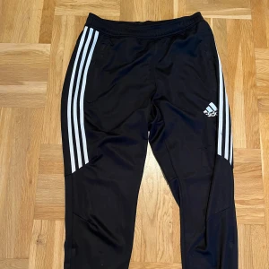 Svarta Adidas träningsbyxor S - Svarta träningsbyxor från Adidas med klassiska vita ränder längs sidorna och logga på låret. Byxorna har elastisk midja och är gjorda i lätt och ventilerande polyester, perfekt för sport eller chill. ClimaCool-teknologi för extra komfort.