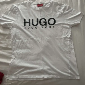 Vit t-shirt från Hugo Boss - Vit t-shirt från Hugo Boss med stor svart HUGO-logga på bröstet. Klassisk rund hals och korta ärmar. Tillverkad i mjuk bomull som känns skön mot huden. Perfekt för dig som gillar stilrena och trendiga plagg med tydlig branding. Pris kan självklart diskuteras 
