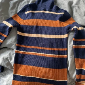 Randig polotröja från Amisu - Snygg ribbad polotröja från Amisu i marinblått, orange och beige. Tröjan har lång ärm, smal passform och höga krage. Perfekt för dig som gillar retrovibbar och vill ha en färgglad look till hösten eller vintern.