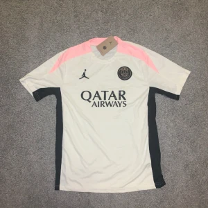 PSG x Jordan vit och svart fotbollströja - Snygg PSG x Jordan fotbollströja i vitt med svarta och rosa detaljer. Tröjan har korta ärmar, svart logga och Paris Saint-Germain-märke på bröstet. Tillverkad i lätt och ventilerande material, perfekt för träning eller match.