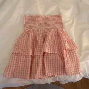 Rosa rutig volangkjol från Kappahl - Supersöt rosa och vit rutig kjol från Kappahl med smock i midjan och dubbla volanger. Perfekt för dig som gillar en feminin och somrig stil. Kjolen är kort och har en luftig känsla, vilket gör den extra bekväm och lätt att bära.