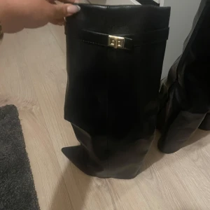Svarta stövlar med spetsig tå - Snygga svarta stövlar i skinn med spetsig tå och dekorativt spänne i guld upptill. Stövlarna har en hög och slouchy passform som ger en cool siluett. Perfekta att styla till både kjol och jeans för en trendig look.