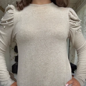 Beige stickad tröja med pärldetaljer - Superfin beige stickad tröja med puffade axlar och pärldetaljer på ärmarna. Långärmad modell med en mjuk och skön känsla, perfekt för dig som gillar en stilren men ändå trendig look.