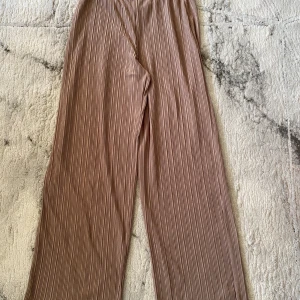 Beige plisserade wide byxor - Säljer ett par snygga plisserade byxor i beige med bred passform och resår i midjan. Byxorna har ett stilrent randigt mönster och är superbekväma, perfekta för en chill och trendig look.