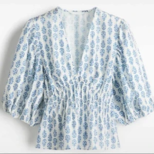 Vit och blå blus med v-ringning - Superfin blus i vitt med blått mönster, v-ringad och med smockad midja. Blusen har puffiga trekvartsärmar och är lätt croppad. Perfekt för dig som gillar en luftig och somrig stil med snygga detaljer.