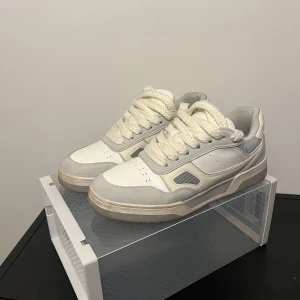 Vita och grå sneakers från Zara - Chunky sneakers från Zara i vitt och ljusgrått med breda, grova snören. Ovandelen är i syntet och mesh, med detaljer i grått och vitt. Platt sula och rund tå ger en clean och modern vibe. Perfekta för dig som gillar stilrena och sportiga skor.