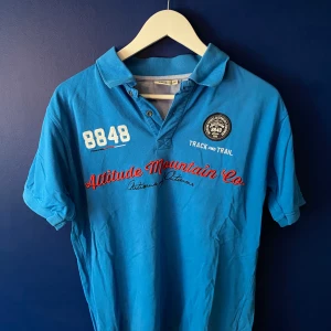 Blå pikétröja från 8848 Altitude - Snygg blå pikétröja från 8848 Altitude med broderade detaljer och tryck framtill. Klassisk krage, korta ärmar och knappar vid halsen. Perfekt för dig som gillar sportig och avslappnad stil.