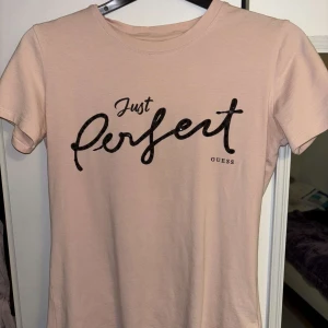 Ljusrosa t-shirt från Guess - Snygg ljusrosa t-shirt från Guess med svart text 'Just Perfect' på bröstet. Klassisk rund halsringning och korta ärmar. Tillverkad i mjuk bomull.