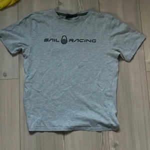 Grå t-shirt från Sail Racing! - Grå t-shirt från Sail Racing med svart logga tryckt över bröstet. Klassisk passform med rund hals och korta ärmar. T-shirten är i mjuk bomull och har en enkel, stilren look som funkar till det mesta.
