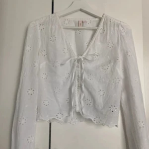 Vit blus från Nelly🌸 - Superfin vit blus med broderade blommor och knyt framtill. Långärmad och croppad modell med v-ringning och vågig kant nertill. Perfekt för dig som gillar romantisk och luftig stil.