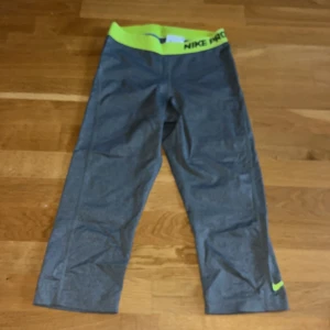 Grå Nike Pro träningstights med neongul midja - Nike Pro träningstights i grått med bred neongul resår i midjan och Nike-logga på benet. Tillverkade i Dri-FIT-material som andas och transporterar bort fukt. Perfekta för gym eller löpning.