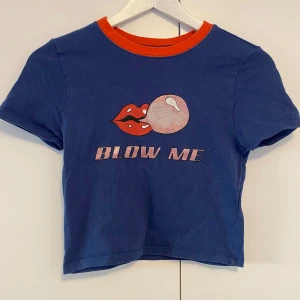 Blå croppad t-shirt från Urban Outfitters - Blå croppad t-shirt från Urban Outfitters med röd-orange ribbad krage. Tryck framtill med röda läppar, bubbelgum och texten 'BLOW ME'. Lite av glittret på trucken har försvunnit. 