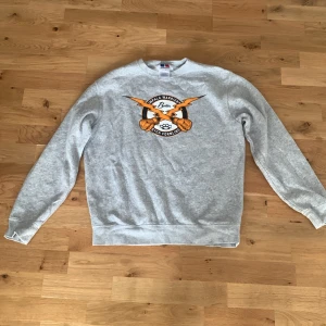 Grå sweatshirt med tryck från Fruit of the Loom - Grå sweatshirt med rund hals och tryck framtill med texten 'Basket Warriors' och basketbollar. Tröjan har ribbade muddar vid ärmslut och nederkant. Perfekt för en sportig look.