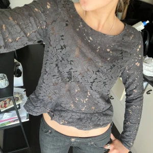 Grå spetsblus med lång ärm - Säljer en grå, långärmad blus i spets med blommigt mönster. Blusen är något croppad och har en rund halsringning. Perfekt för dig som gillar detaljer och vill ha en unik topp i garderoben.