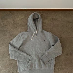 Grå hoodie från Polo Ralph Lauren - Säljer en klassisk grå hoodie från Polo Ralph Lauren med broderad logga på bröstet. Tröjan har huva med vita snören och en stor magficka. Perfekt för en avslappnad stil. Säljs eftersom den e för liten för mig.