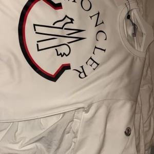 Vit sweatshirt från Moncler - Säljer en stilren vit sweatshirt från Moncler med stort tryck på framsidan i svart och rött. Tröjan har långa ärmar och en rund halsringning. Perfekt för en casual look med en touch av lyx.