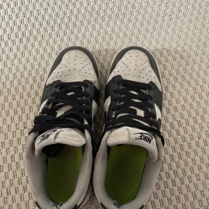 Nike Dunk Low svartvita sneakers - Säljer ett par klassiska Nike Dunk Low sneakers i svart och vitt. Skorna har svart swoosh, svart snörning och vit ovandel med perforerade detaljer. Låg modell med platt sula och gröna innersulor. Perfekta för dig som gillar streetwear.