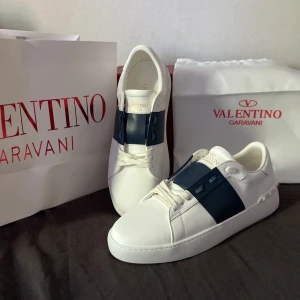Valentino Open sneakers med blå detalj - Säljer ett par vita Valentino Open sneakers med bred blå rem över mitten och ikoniska nitar på hälen. Skorna har rund tå, platt sula och snörning. Perfekta för dig som gillar stilrena och exklusiva sneakers.