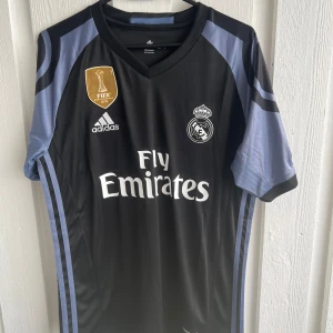 Real Madrid fotbollströja Ronaldo 7 - Snygg svart Real Madrid fotbollströja med Ronaldo 7 på ryggen. Tröjan har korta ärmar, grå detaljer och klubbmärke samt sponsortryck på bröstet. Adidas-logga och FIFA-märke framtill. Perfekt för fans!
