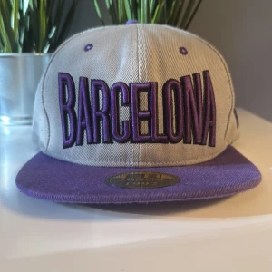 Grå och lila keps med Barcelona-tryck nypris 800 knappt andvänd - Snygg keps i beige med lila detaljer och stor broderad text 'Barcelona' framtill. Lila skärm och justerbar snapback baktill. Broderad logga på sidan och baksidan. Perfekt för dig som gillar streetstyle.