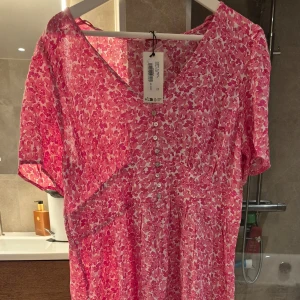 Rosa blommig klänning från Indiska - Säljer en rosa blommig klänning från Indiska i storlek L. Klänningen har v-ringning, korta ärmar, knappar framtill och en slits nertill. Materialet är lätt och luftigt, perfekt för varmare dagar. Helt ny. Hund finns i hemmet.