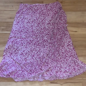 Rosa blommig omlottkjol - Säljer en rosa omlottkjol med vitt blommönster. Kjolen har knytband i midjan och är tillverkad i ett lätt och luftigt material, perfekt för varmare dagar. Den har en mjuk och flowy känsla och passar dig som gillar en feminin stil.