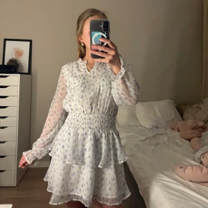 Vit blommig klänning från Gina Tricot - Supersöt vit klänning från Gina young med små blå blommor. Klänningen har långärmade transparenta ärmar, smock i midjan och volangdetaljer på kjolen. Använd 1 gång💞