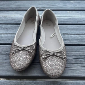 Glittriga ballerinaskor med rosett - Säljer ett par glittriga ballerinaskor i beige med små rosetter framtill. Skorna har rund tå och är täckta av små paljetter som ger en snygg och festlig look. Perfekta för dig som vill sticka ut lite extra!