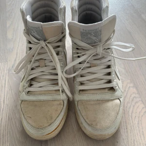 Vita höga sneakers från Hummel - Säljer ett par vita höga sneakers från Hummel med klassisk design och snörning framtill. Skorna har gummisula och är tillverkade i textil och syntetmaterial. Storlek 38 bra skick förutom att dom är smutsiga 