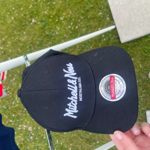 Svart keps från Mitchell & Ness - Svart keps från Mitchell & Ness med vit broderad logga framtill och klassisk böjd skärm. Justerbar passform baktill. Perfekt accessoar för en sportig och avslappnad stil.