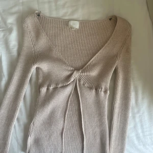Beige ribbad topp med knyt från H&M - Säljer en beige ribbad långärmad topp från H&M med v-ringning och knytdetalj framtill. Toppen är figurnära och har en mjuk, stretchig känsla. Perfekt för lager-på-lager eller som den är.