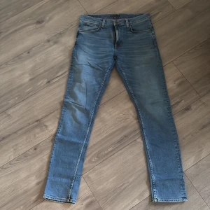 Blå Lean Dean jeans från Nudie Jeans - Säljer ett par klassiska blå Lean Dean jeans från Nudie Jeans. Modellen har smal passform, fem fickor och orangea sömmar. Tillverkade i 99% bomull och 1% elastan för lite stretch. Perfekta för dig som gillar en tidlös jeanslook.