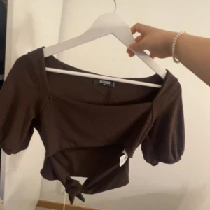 Brun croppad topp från Missguided - Säljer en brun croppad topp från Missguided med korta puffärmar och knytdetalj framtill. Toppen har en fyrkantig ringning och är tillverkad i ett ribbat material som ger en snygg struktur.